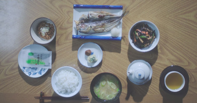 un desayuno tradicional japones, la caballa en un plato cuadrado, rodeada de platos redondos, sopa, té, arroz,  ensalada, un plato de habas, condimentos, encuertidos...