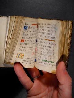 A photograph of a hand holding a small book open. The pages are handwritten with decorative initials.