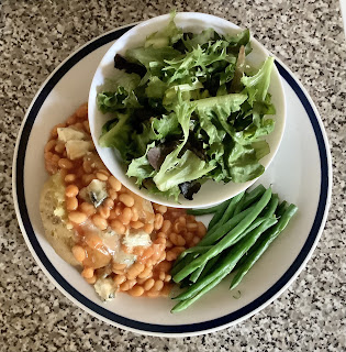 Plate with a jacket potato topped with baked beans and some bits of blue cheese, with some green beans on the side and a bowl of salad leaves