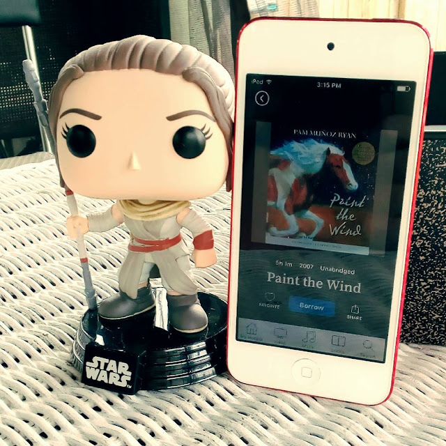 A large-headed Funko Pop bobblehead of Rey from Star Wars stands outdoors beside a white iPod with Paint the Wind's cover on it. The cover features a galloping white and chestnut paint horse.