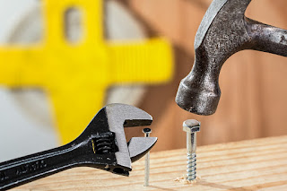 Image shows a piece of wood with a nail and a bolt part-way into the wood. A hammer is held over the nut, and a spanner is held over the nail.