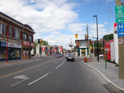 Looking along a street at a major urban intersection with red sidewalks and red concrete crosswalks on all corners except on the right, where there are grey unit pavers. (Somerset Street West looking east across Bronson Avenue)