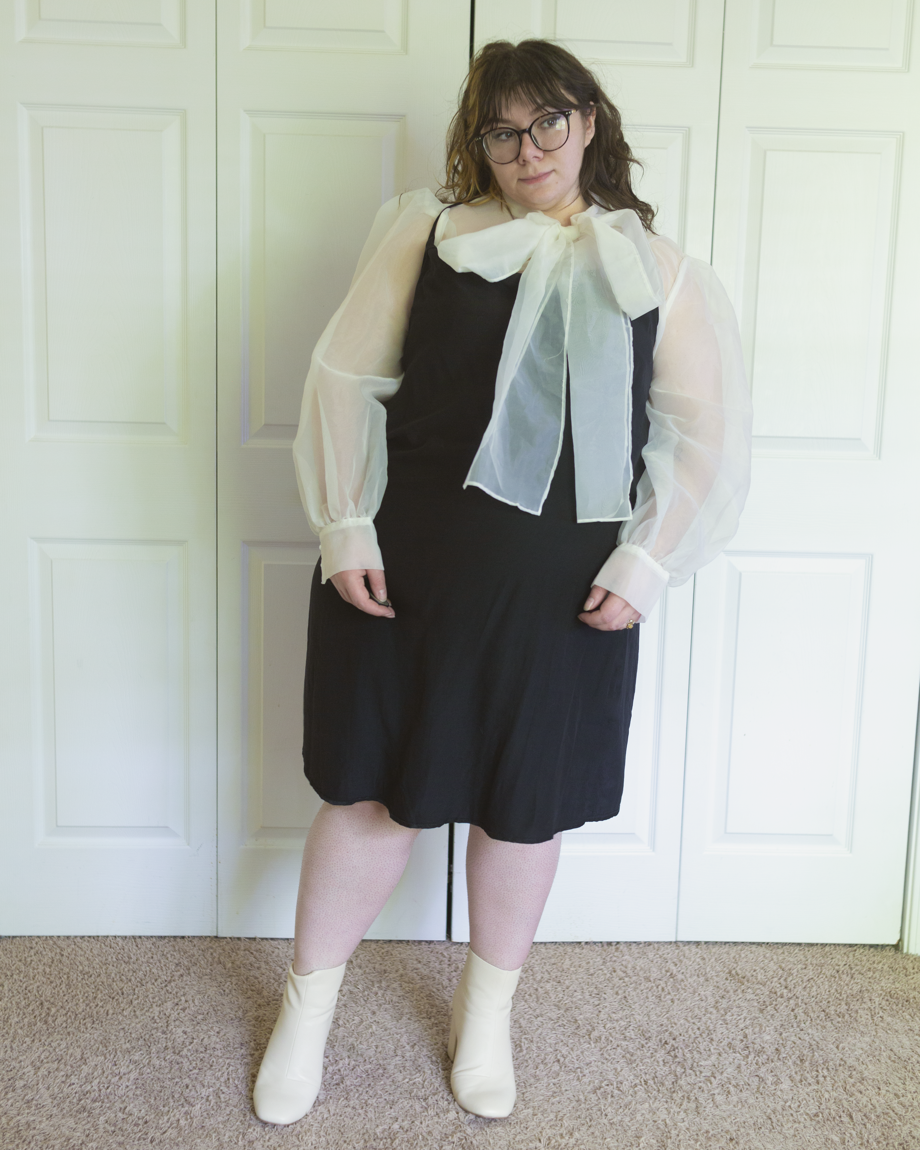 An outfit consisting of a white sheer bishop sleeve blouse with an attached oversized pussy bow, tied in a bow around the neck under a black satin slip dress and cream midi boots.