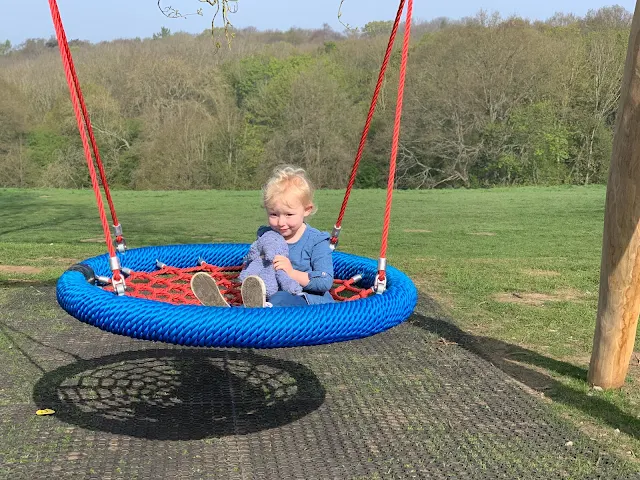 A toddler on a spider web swing with trees in the background