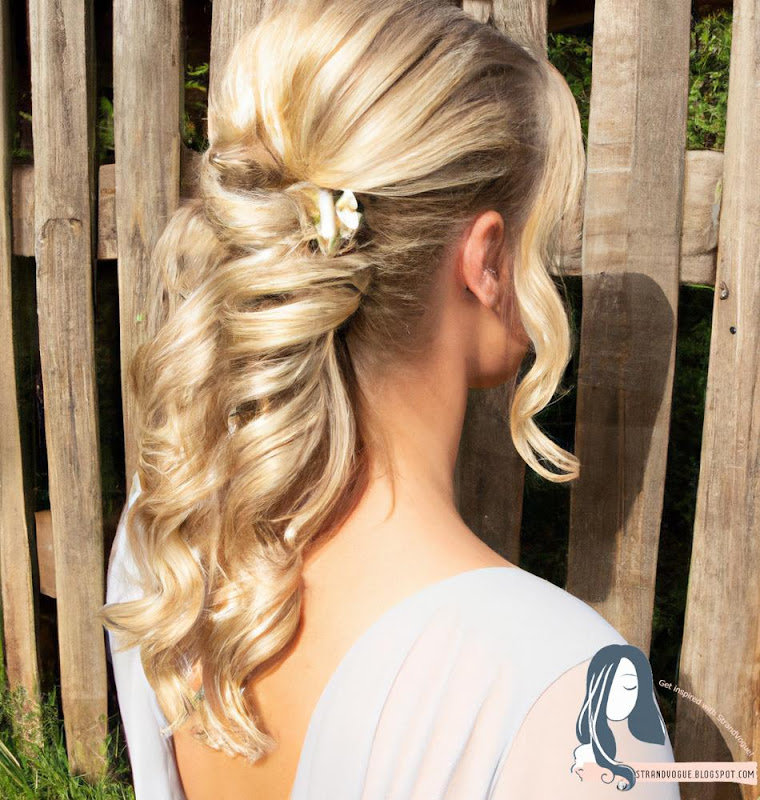 Image of Half-Up Twist and Pin: A woman with blonde hair is standing in front of a wooden fence. Her hair is styled in a half-up twist and pin, with loose curls cascading down her back.