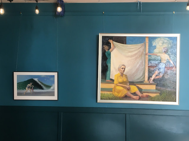 Peacock blue gallery wall with pastel painting of mother applying sunscreen to her daughter's nose as a large wave breaks in the background, and an oil painting of three women, a mother, daughter, and grandmother on a porch overlooking a town with a thundercloud on the horizon.