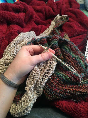 A white hand holding gold circular knitting needles up, in the middle of a row of white lace attached to a crumpled pile of teal, pink, gold, and green garter stitch. The lace-edged part of the shawl stretches out and away on top of a red fleecy blanket.