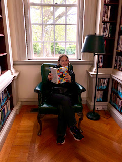 Natalie is making herself comfortable in a reading chair by a window, pretending to read a book by Louise Erdrich.