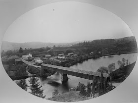 A black and white photograph of a long covered bridge over a river. A small cluster of buildings is on the opposite side.