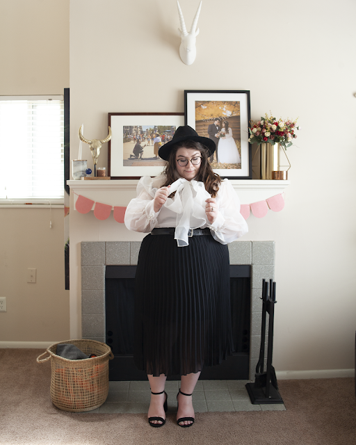 An outfit consisting of wide brim black fedora, sheer whit blouse with an attached bow tucked into black pleated midi skirt and black ankle strap heels.