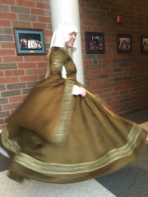 A young white woman spinning to her left in a red brick hallway with large checkerboard tiles, wearing a sheer white veil and green-gold silk dress with pendant sleeves lined in peach and a full skirt. The collar, sleeves and skirt hems, and upper arms are decorated with bands of gold floral embroidery on brown, and she has a long gold belt. She's side-on to the photographer, grinning, with her skirt and near sleeve completely belled out by her motion and her white chemise showing through the lacing gap at her side.