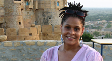Jennifer James smiled at the camera as stands before a large stone and adobe facade, with a wide valley in the background. She has short dread locks pulled into a high ponytail, and her sunglasses pushed up on her head. She is wearing large, silver earrings, and a pink v-neck cotton blouse.