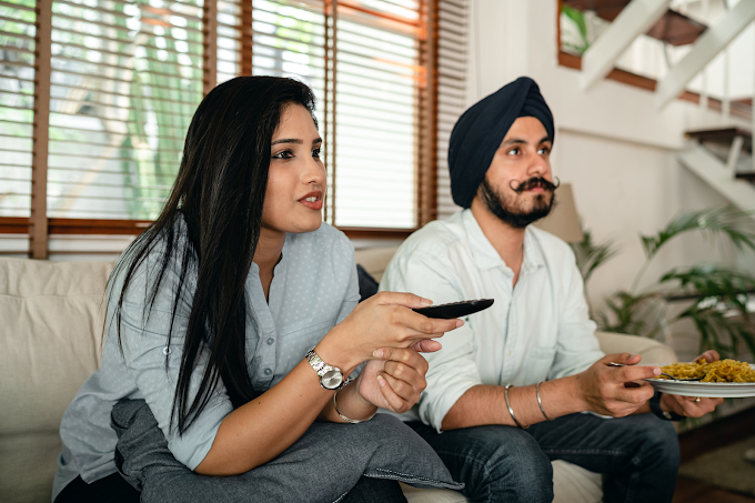 Homem e mulher sentados no mesmo sofá vendo TV. Mulher está com controle na mão enquanto o homem come macarrão