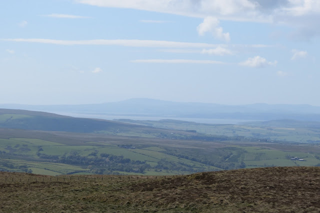 A view across Lancashire farmland to the waters of Morecambe Bay, with hazy outlines of Lake District fells beyond it.