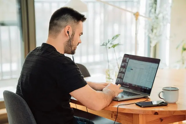 Hombre con playera negra, sentado en una silla gris, trabajando en una macbook que está sobre una mesa de madera