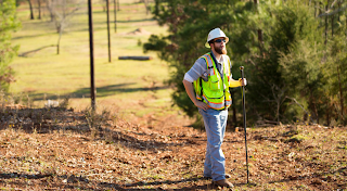 man wearing hard hat and yellow vest standing on a landscape with trees and poles holding electricity cables