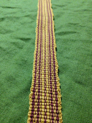 A length of striped rose and gold plain inkle weaving with slightly wobbly selvedges, laid out on green fabric.