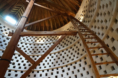 Interior of a dovecote: the walls are white, with a checkerboard pattern of square niches for the pigeone; a central wooden post extens to the roof, with a ladder which is curved to match the curve of the dovecote.