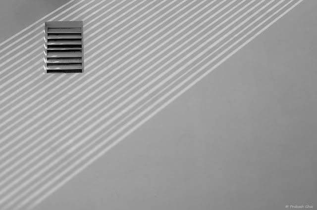 A Black and White Minimal Art Photograph of a Window with Horizontal Blinds, placed on a White Wall with Diagonal lines created by Light and Shadows
