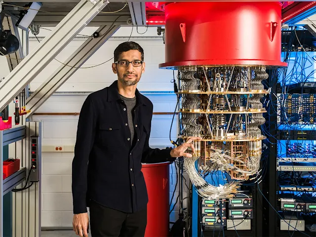 Google CEO Sundar Pichai stands in front of a quantum computer. He wears a black suit and smiles as he gestures toward the complex machinery.