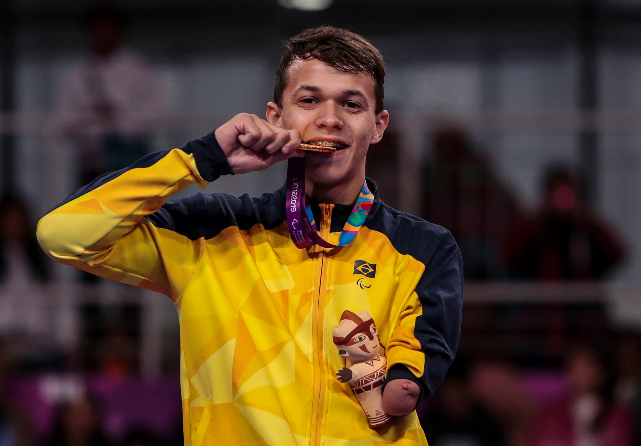 Nathan Torquato sorri ao morder a medalha de ouro e segura mascote