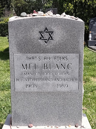 Photo of a gravestone; gray, upright granite, engraved Jewish star. Text  reads "That's all, folks. Mel Blanc Man of 1000 Voices Beloved husband and father. 1908-1989." The gravestone has small stones on it that people have left - a Jewish tradition.