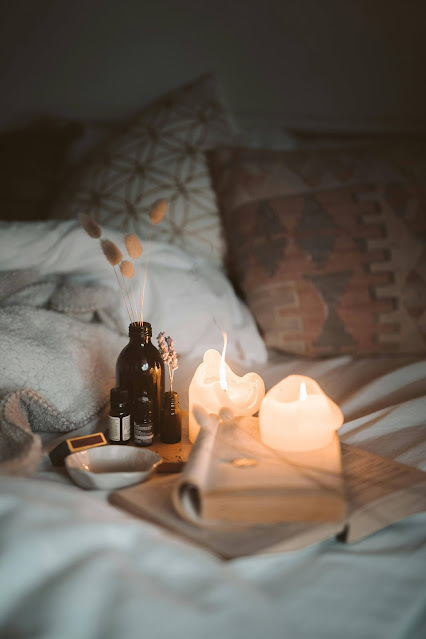 Reed diffuser, essential oils, lighted candles, porcelain glass dish, open books in cozy bed with bedsheets, cover & pillows.