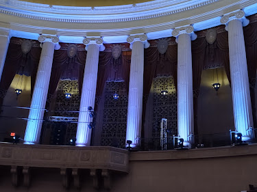 Photo. The back of a large, circular auditorium with white columns two stories tall.