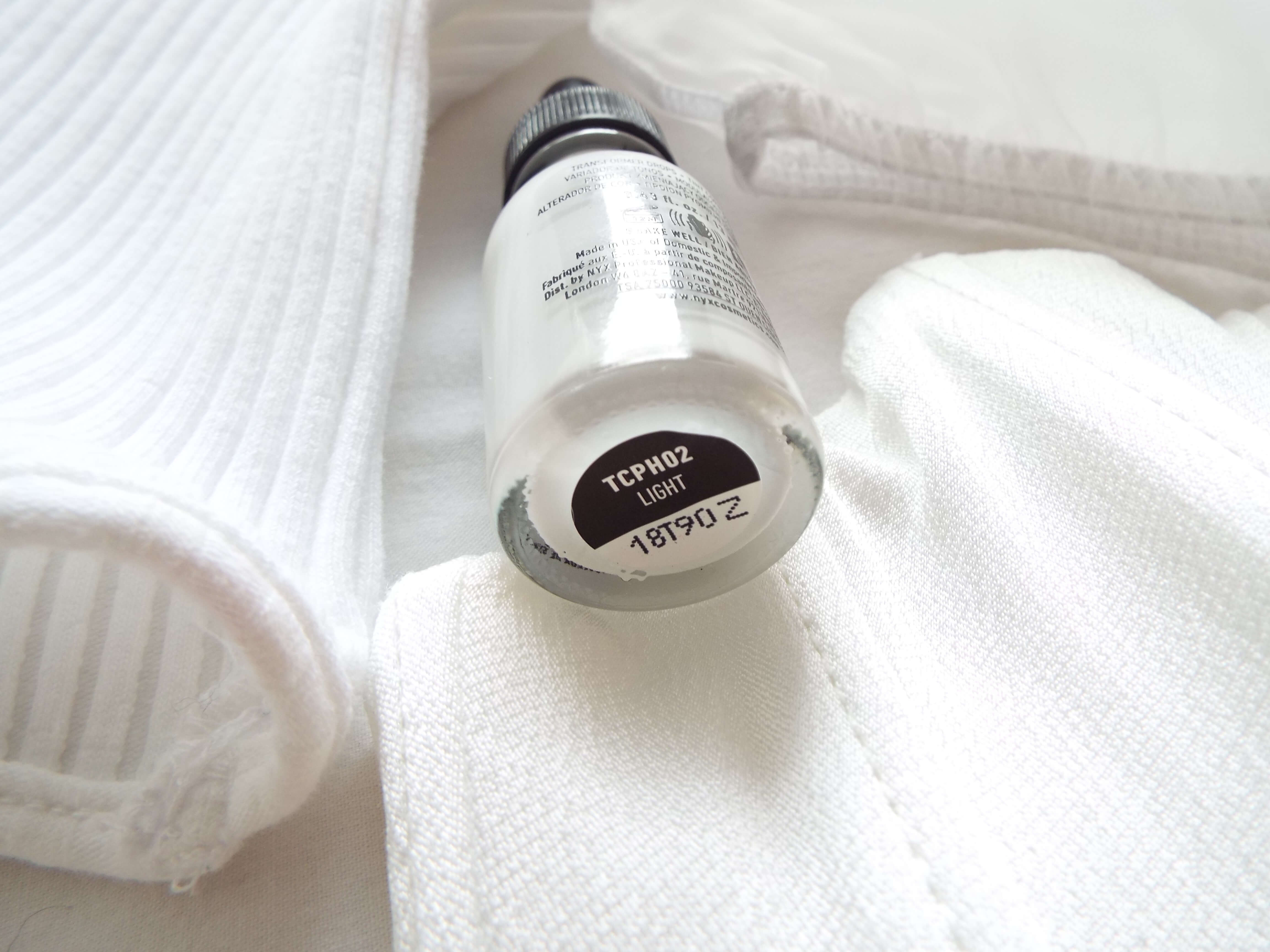 Glass dropped bottle of the NYX Total Control Pro Hue Shifter lightening drops, between clothes on white bedding.