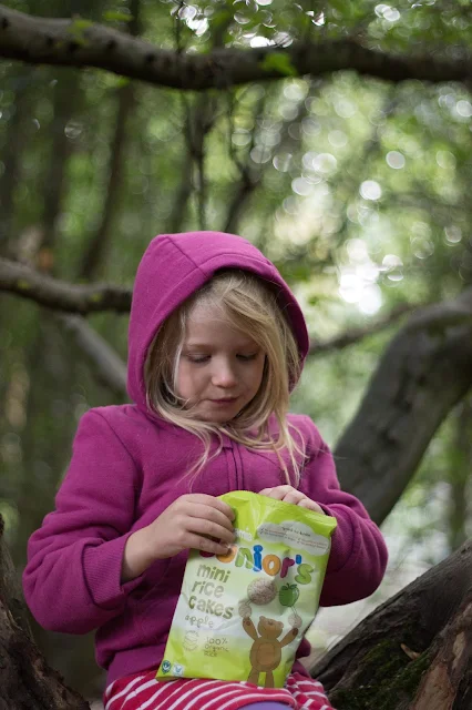 A girl in a pink hoody sits in a tree with a bag of mini rice cakes