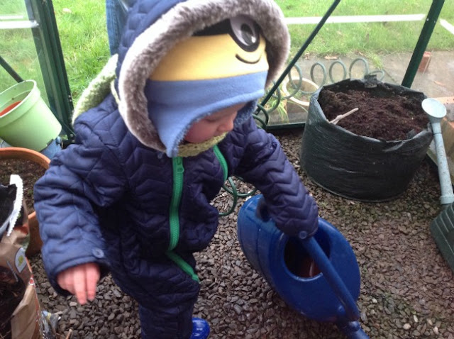 toddler with watering can in green house all wrapped up with scarf and hood