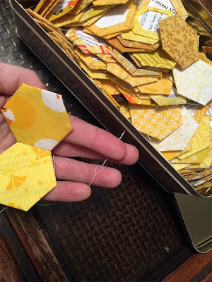 A white hand cradling two yellow fabric hexagons seamed together, with a small needle pinned between outstretched fingers, next to a shallow rectangular tin full of basted yellow hexagons.