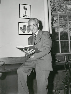 A black and white photograph of an older man in a suit, leaning against a desk with an open book in his hand.