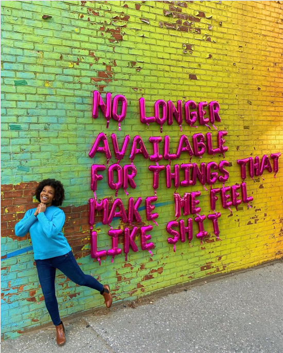 Picture of a Black woman standing on one leg, hands folded, smiling, in front of a wall letters saying NO LONGER AVAILABLE FOR THINGS THAT MAKE ME FEEL LIKE SHIT