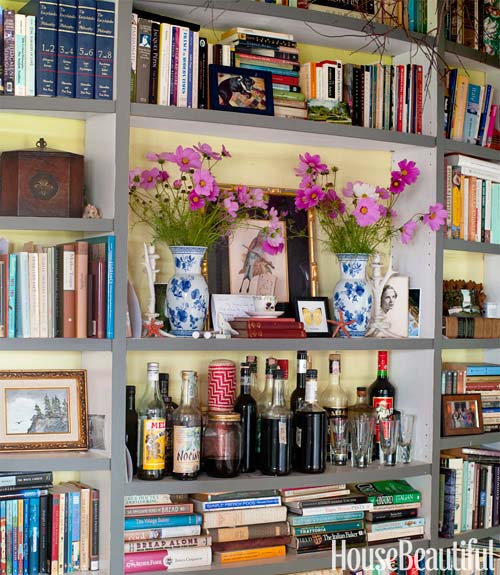 house beautiful's built in bookshelves stuffed full of books, larger shelves hold items like flower arrangements, a small bar and other trinkets