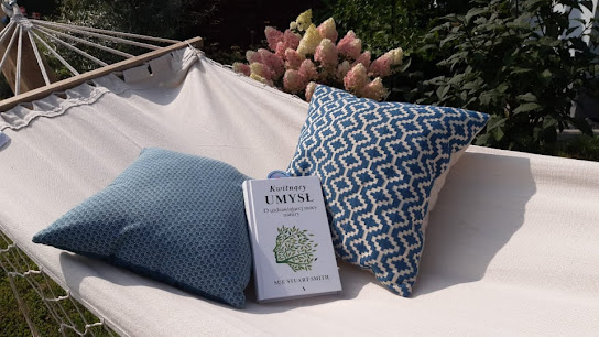 W ogrodzie na białym hamaku leżą dwie poduszki w biało-niebieskie wzorki oraz książka "Kwitnący umysł. O uzdrawiającej mocy natury" Sue Stuart Smith.