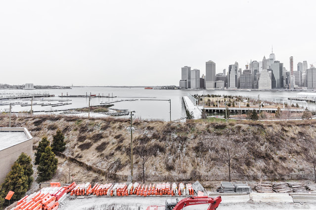Overcast view of Lower Manhattan's skyline from Brooklyn Heights, with barren trees and a grassy slope in the foreground. The East River lies between, with idle boat docks and a solitary red tugboat. Construction barriers and machinery suggest ongoing development. Despite the gray skies, the city's iconic skyscrapers stand resilient, showcasing the quiet yet persistent energy of a metropolis in winter.