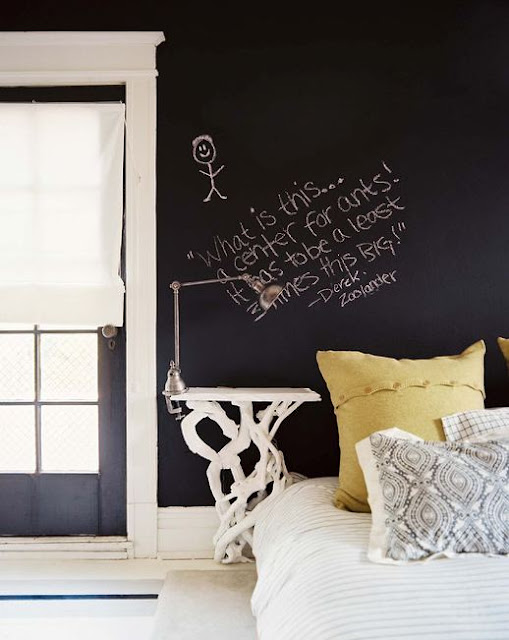 Bedroom with French doors with Roman shades, a white nightstand designed in the shape of branches holds a silver clamp light and instead of a headboard, the white bed with green and paisley accent pillows, has a black chalkboard