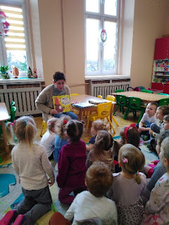 tło : kolorowa sala przedszkolna, pani czyta i pokazuje ilustracje , dzieci siedzą na podłodze i słuchają z zainteresowaniem zajęć.