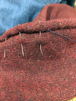 A close-up of the rolled edge of seam allowance, pinned at the left, with a strand of navy-blue thread emerging from the fold at the right. The weave of the garnet-red cloth is just visible through a fine layer of fuzziness on the surface.
