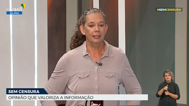 Ana Moser é morena e está com uma camiseta em tons terrosos (bege) e seu cabelo (tons brancos) está preso. Ela está no estúdio da TV Brasil com fundo branco e leds azuis