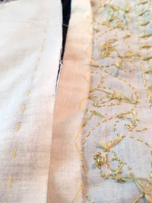 Two piece of white fabric abutting each other; the right piece has the hem folded back and tacked down with yellow thread, and shows the messy back of an embroidered panel. The left piece is basted in place with yellow thread, but otherwise empty.