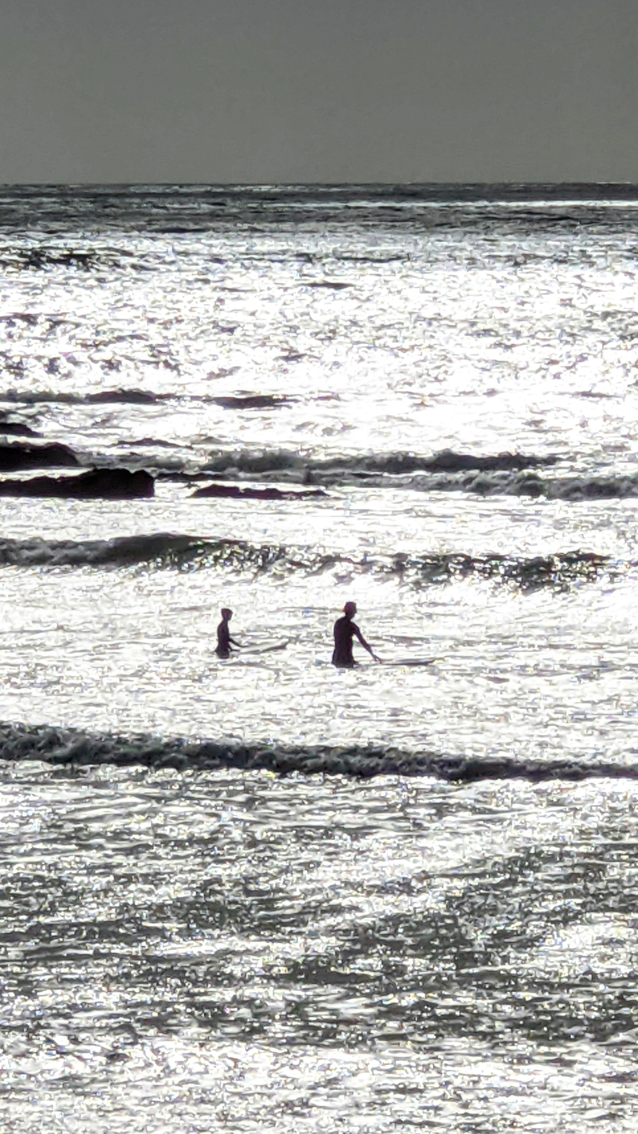 Monochrome view of surfers paddling out to the waves on a sun reflected sea