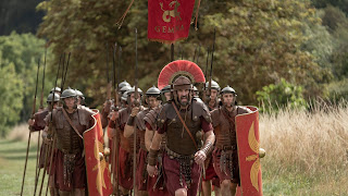 A group of Roman legionaries determinedly marching up a hill, led by Lee Mack as a centurion. The Fifteenth Doctor is somewhere at the back.