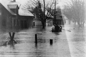 A black and white photograph of a flooded street. A few figures are seated in a boat, paddling through the water.