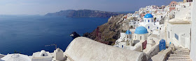 A panoramic photo of the city of Iola, with its characteristic whitewashed buildings, several of which are topped by blue domes, on the craggy island of Santorini, with the Mediterranean Sea visible in most of the left half of the photo.