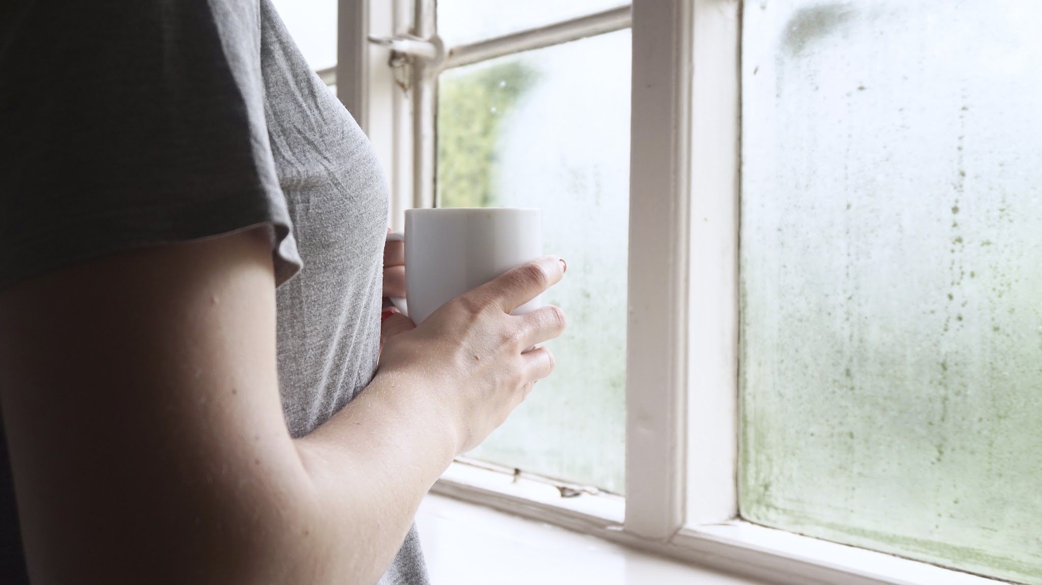 woman looking out of steamed up window with cup of tea in hands