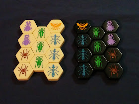The twenty-two pieces from the base set: the eleven white pieces on the right and the eleven black on the left. Each set is a thick Bakelite hexagonal tile with an arthropod embossed into the top and paint poured into the recessed area. On each side, there are two purple beetles, two brown garden spiders, three green grasshoppers, three blue worker ants, and one queen bee.