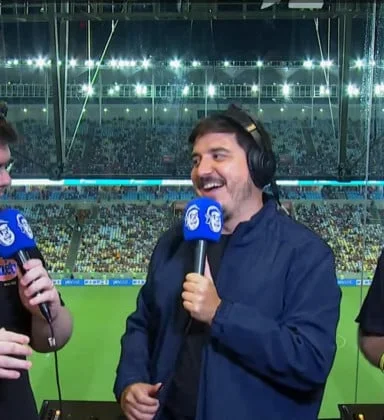 Luis está de casaco e com o microfone da Cazé Tv, que é azul. Ele usa fone e está de pé na cabine com o Maracanã ao fundo