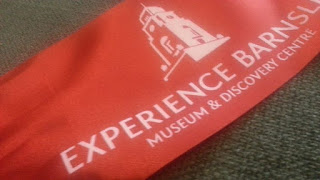A broad red silky ribbon with the Barnsley Town Hall logo and the words Experience Barnsley Museum and Discovery Centre